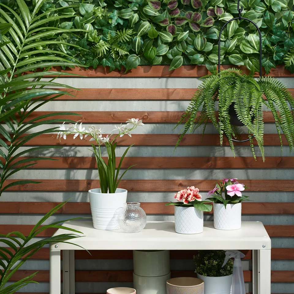 FEJKA artificial plant wall with flowers and greenery against wooden slats, plus potted plants on table.