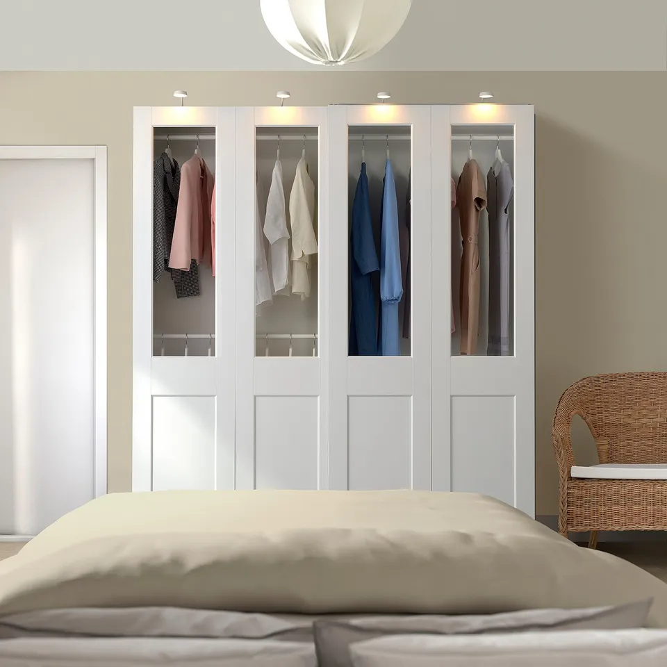 Modern bedroom with white PAX wardrobe, glass doors, lit clothes, and wicker chair.