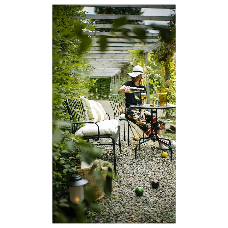 Person in garden, sitting at table with LÄCKÖ black metal chairs, wearing hat, drinking from glass, surrounded by green plants.