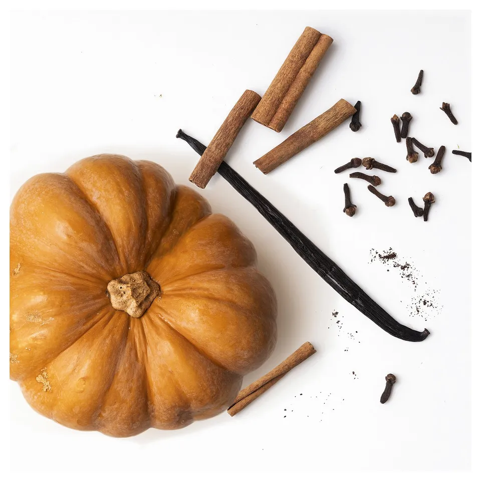 Pumpkin-shaped yellow candle with cinnamon, cloves, and vanilla beans spices scattered around.