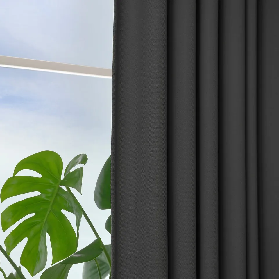 Dark grey KORGMOTT block-out curtains hang by a window, allowing light control and privacy. A green plant sits beside them.