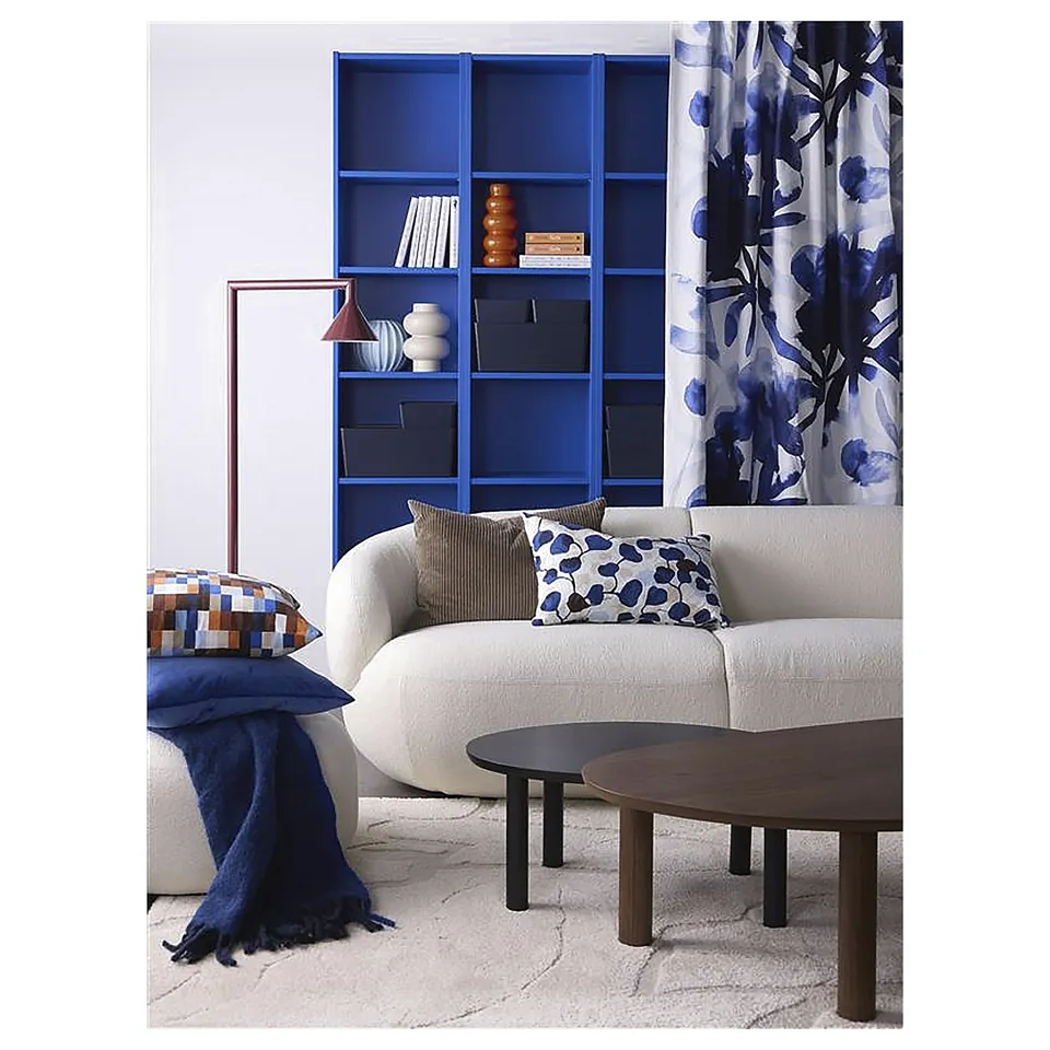 Modern living room with white couch, blue accents, patterned pillows, wooden tables, blue shelf, and floral curtain.