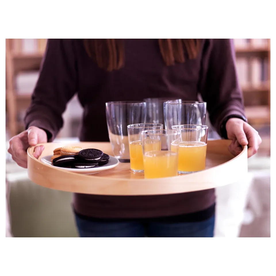 Person carrying SKALA birch tray with orange juice and cookies, clear glass cups in light, casual setting.