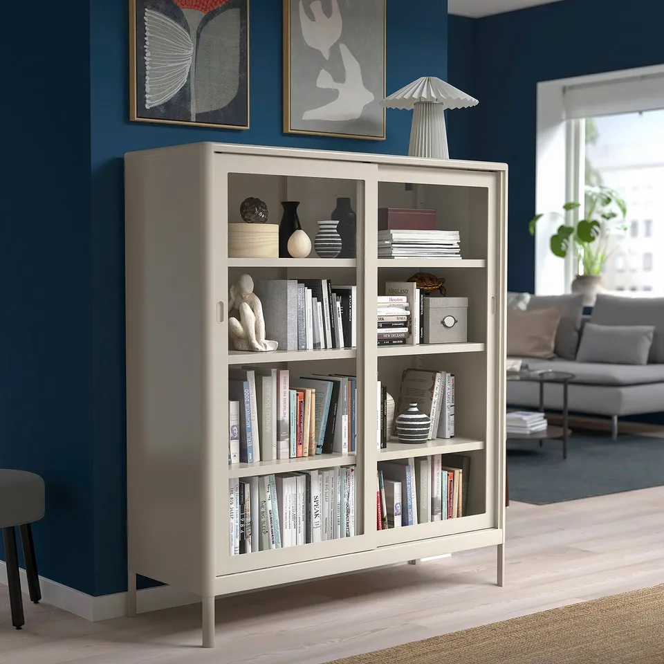 The IDÅSEN bookshelf in white with glass doors stands tall, holding books & decor. Its larger than the stool & sofa in a blue-walled room.