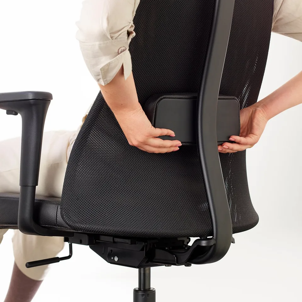 Person adjusting lumbar support on black office chair with mesh back and armrests.