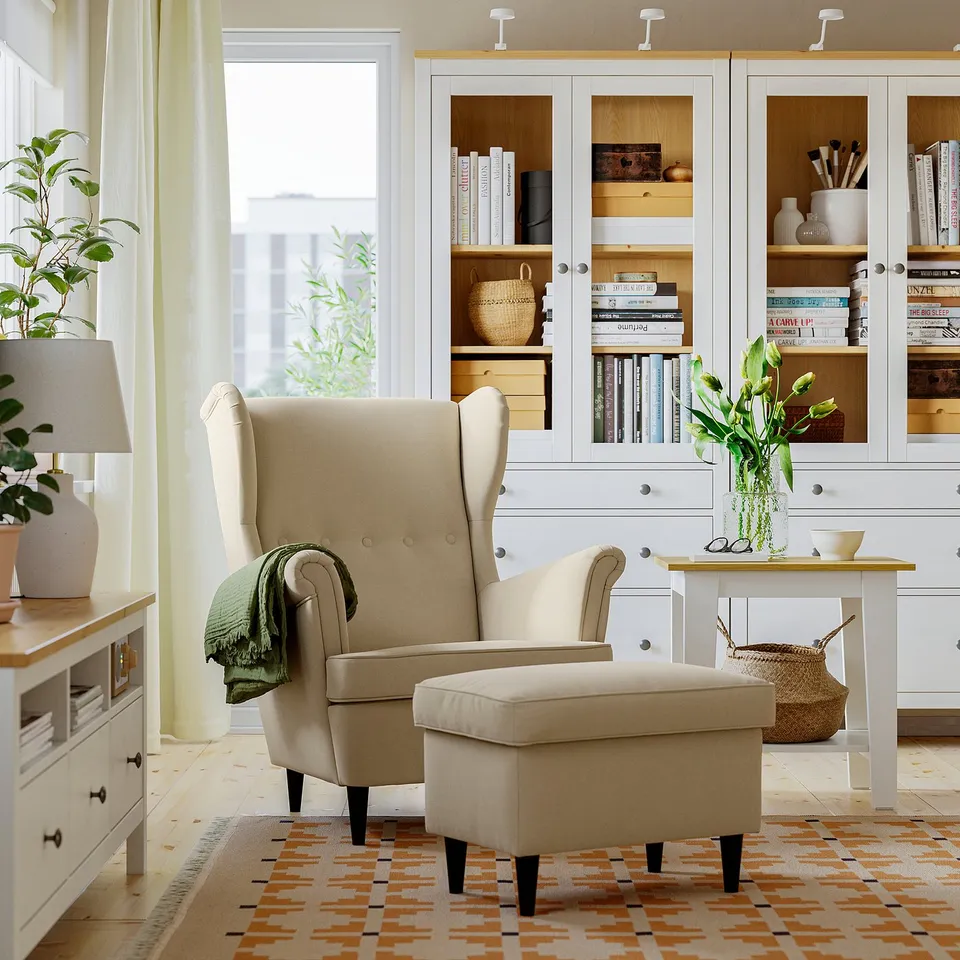 Beige STRANDMON wing chair & ottoman in cosy living room, with bookshelf, green blanket, & side table with flowers.