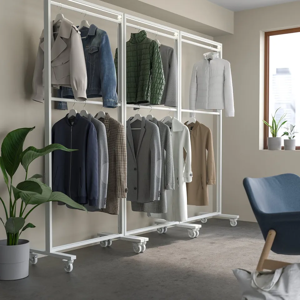 Double MITTZON coat racks, white steel, with wheels, holding various clothes and jackets, positioned near a large potted plant and a blue chair.