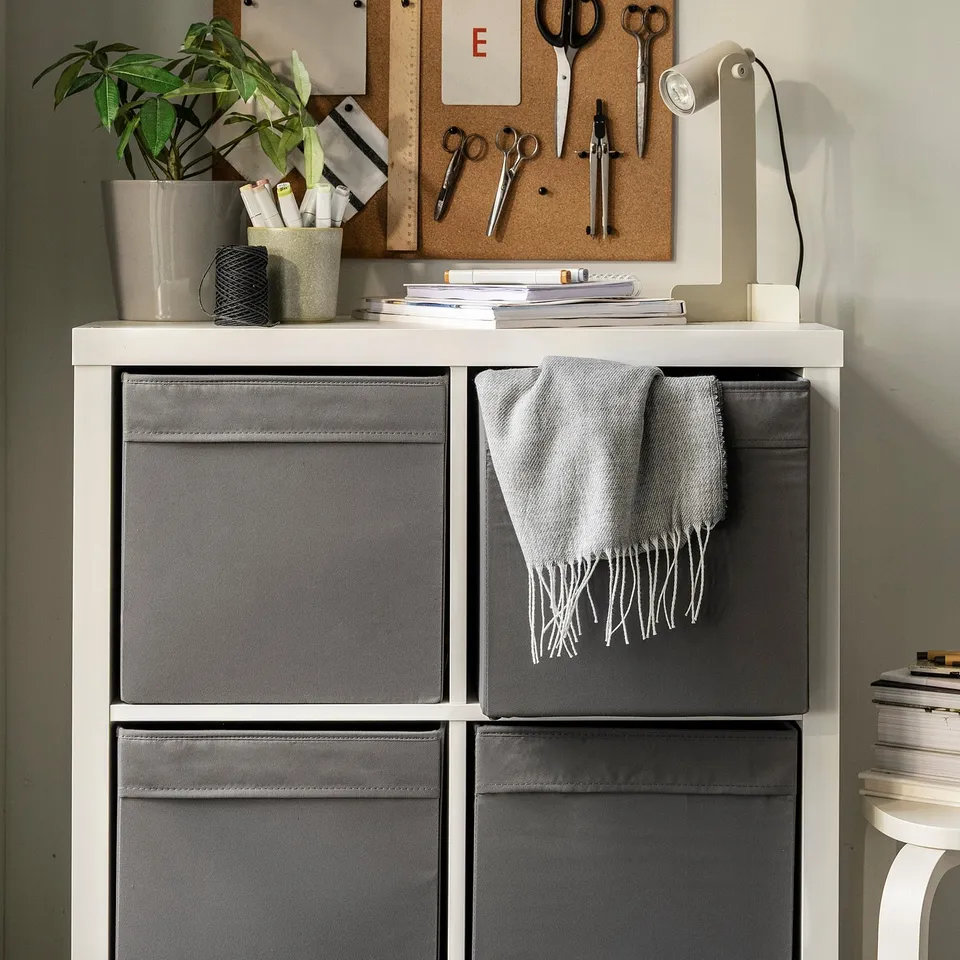 White shelving with 4 grey DRÖNA boxes, cloth in one, plant, notebooks, cork board with notes.