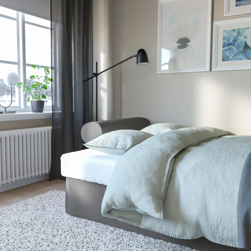 Minimalist bedroom with large bed, light blue blanket, and wall lamp. A plant by the window and white frames decorate the room.