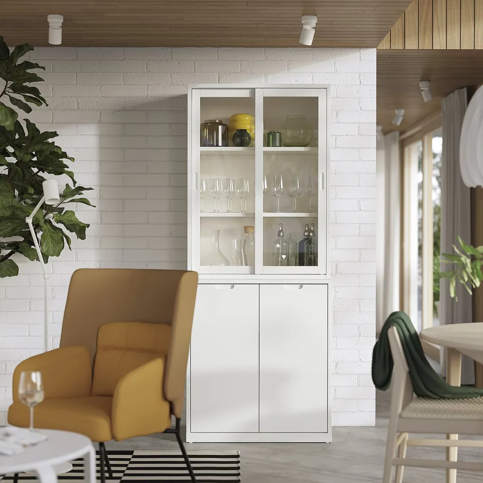 Modern living room with TONSTAD off-white cabinet displaying glassware, flanked by a yellow chair and a large plant.