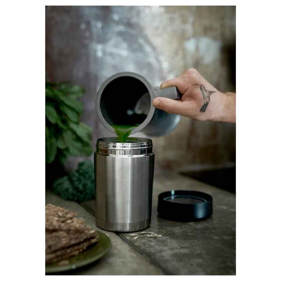 Person pouring green liquid into EFTERFRÅGAD stainless steel thermos. Dark countertop with plate of food beside it.