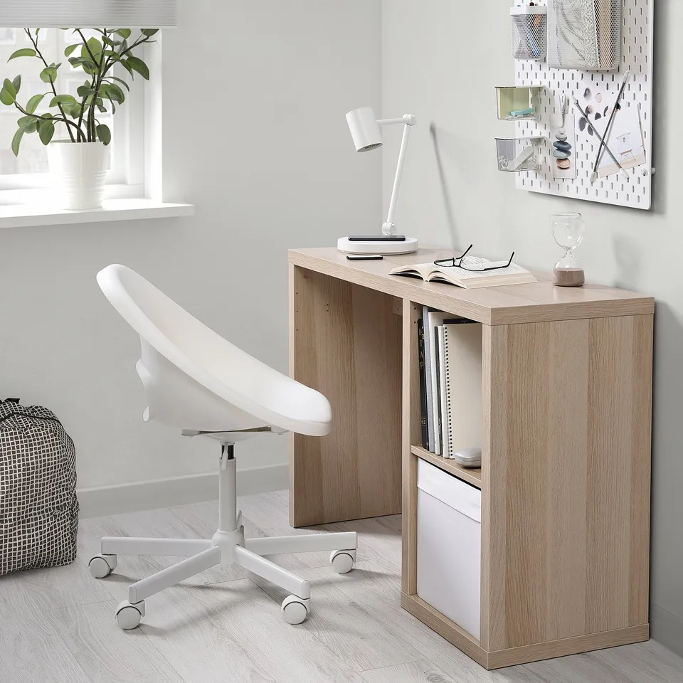Minimalist home office setup with a light brown KALLAX desk next to a window, featuring a white swivel chair, lamp, and decorative items.