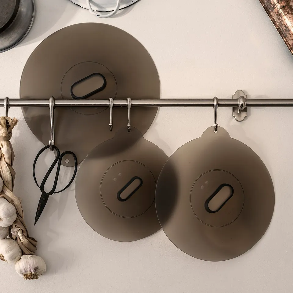 Three grey silicone lids hang from hooks on a metal bar, showcasing their circular shape and vacuum seal design. Garlic and scissors are nearby.