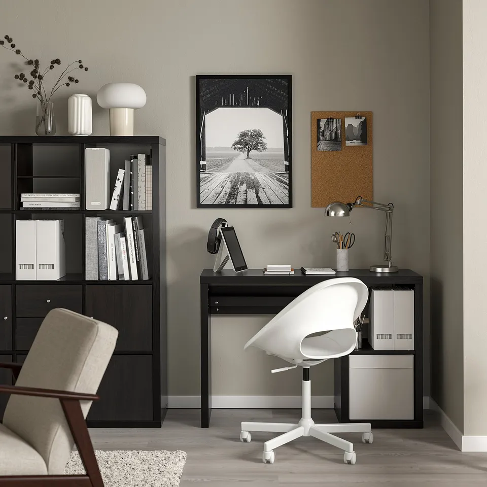 Minimalist workspace with black KALLAX bookshelf, white books, black desk, white chair & lamp. Black photo frame above.