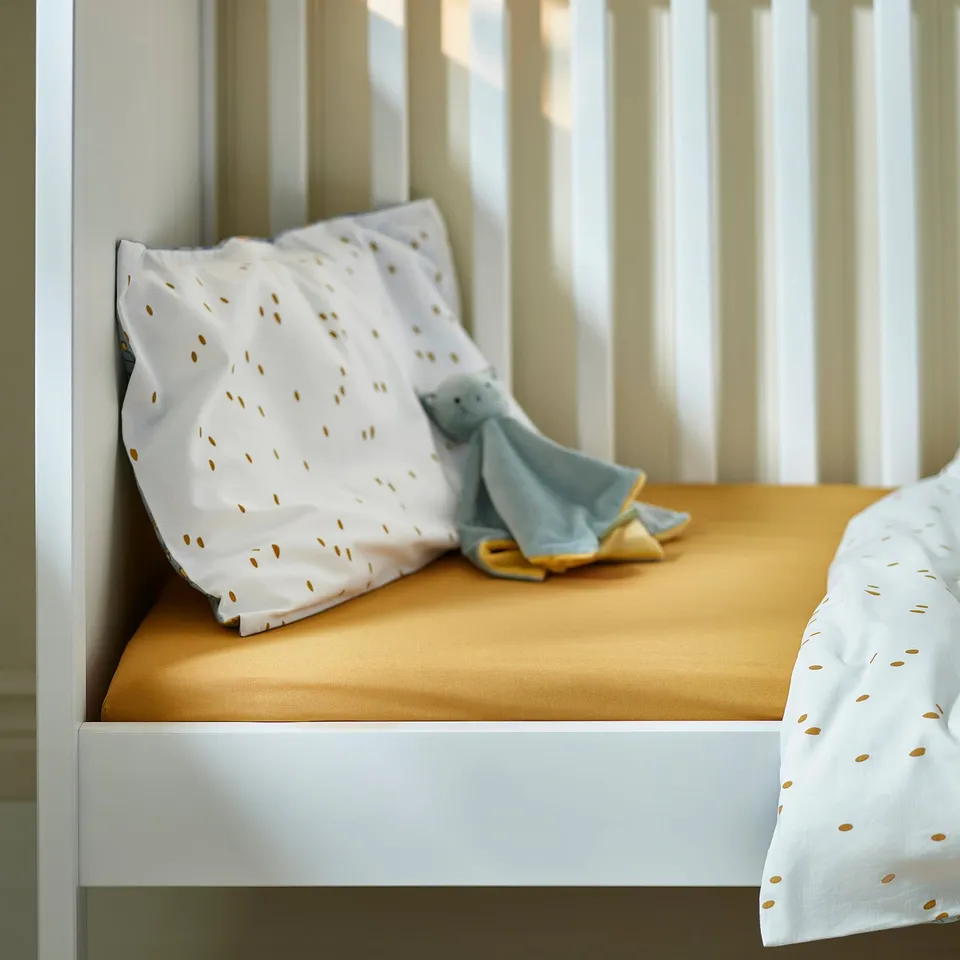 A white cot with a mustard fitted sheet, white pillow with beige dots, and a blue cuddly toy.