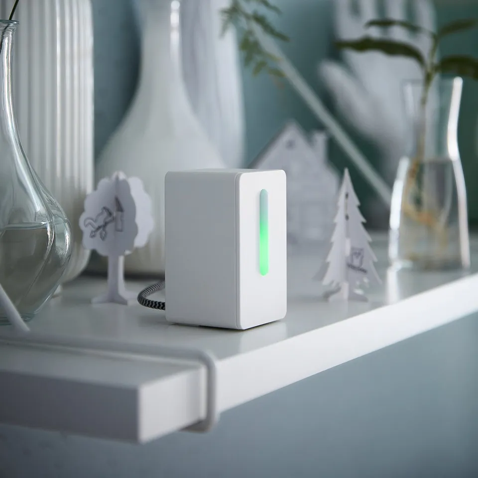 Small white device glows green on shelf with vases, decorative trees, and plants.