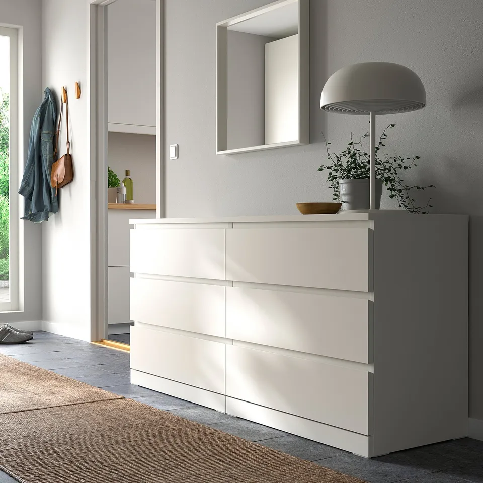 Modern MALM dresser with smooth drawers. Blue jacket and purse hang nearby. Wooden rug and grey lamp in room.