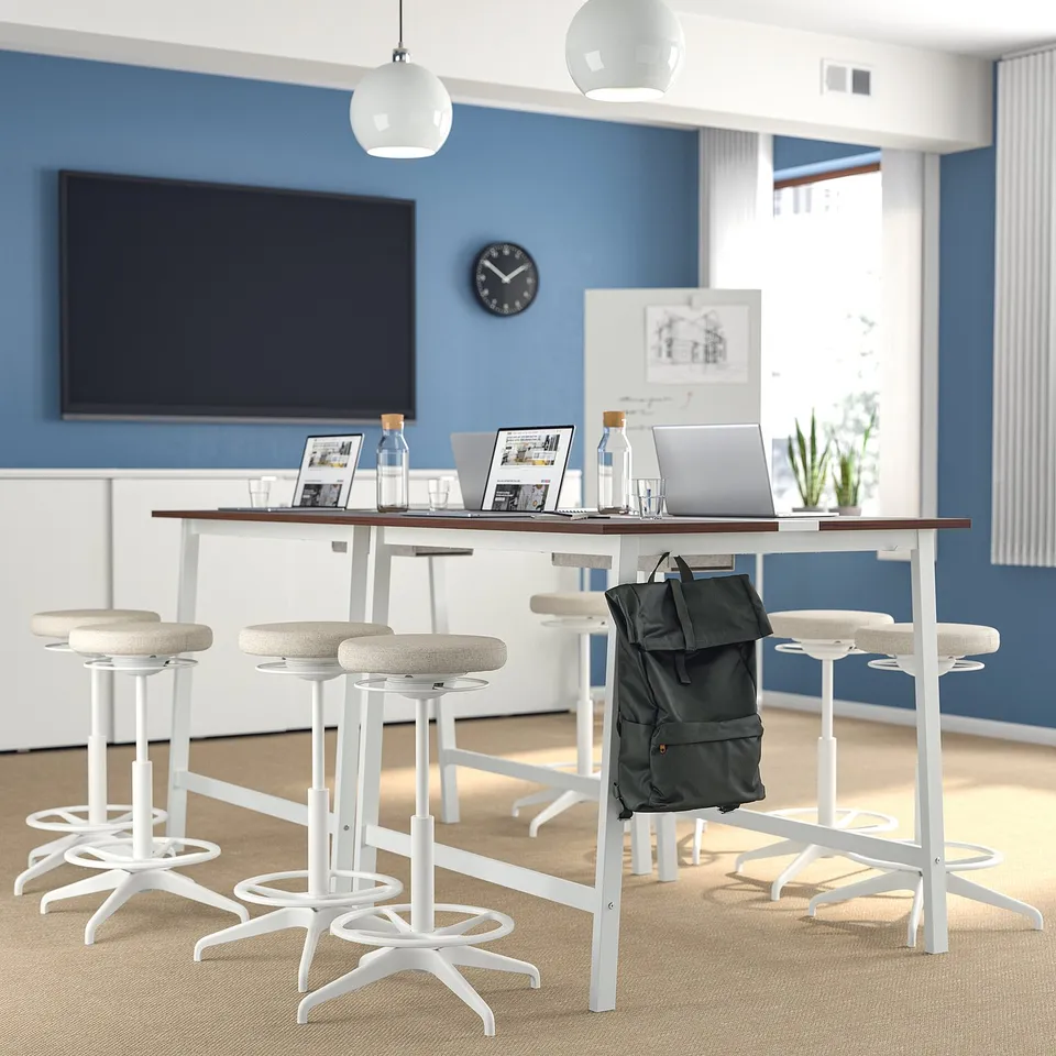 MITTZON conference table with stools, backpack, laptops, bottles, and frames. Blue wall with tv and clock. Minimal, professional decor.