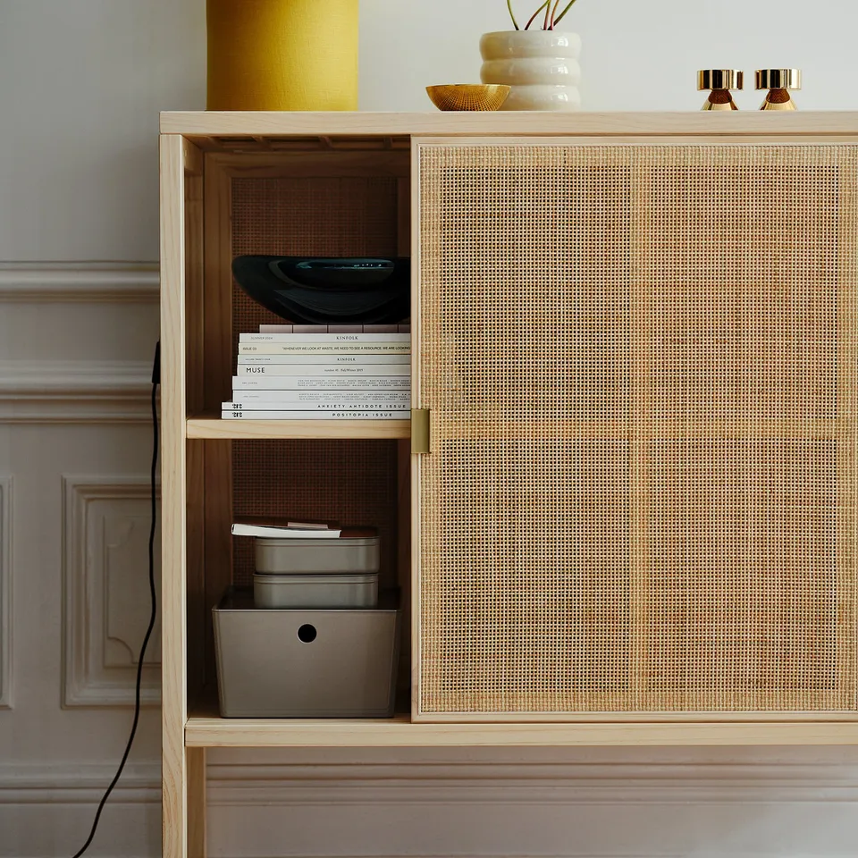 Stylish rattan cabinet with brass handles and sliding doors, adjustable feet, and open storage with books and containers.