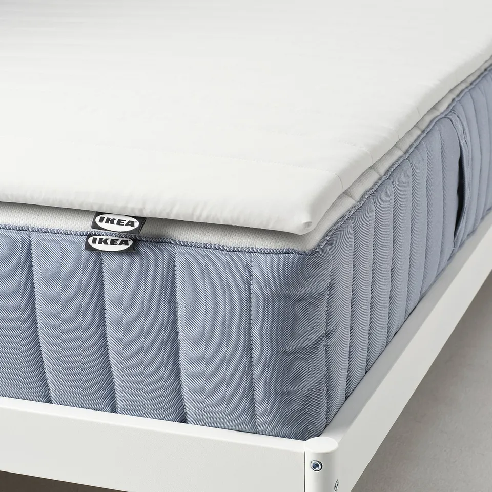 A grey mattress pad with IKEA tags placed on a white bed frame, offering comfort and support with a soft, smooth cover.