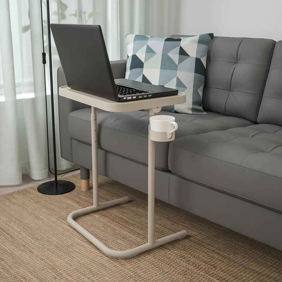 Beige BJÖRKÅSEN laptop stand next to grey sofa, holding laptop & cup, portable & adjustable.