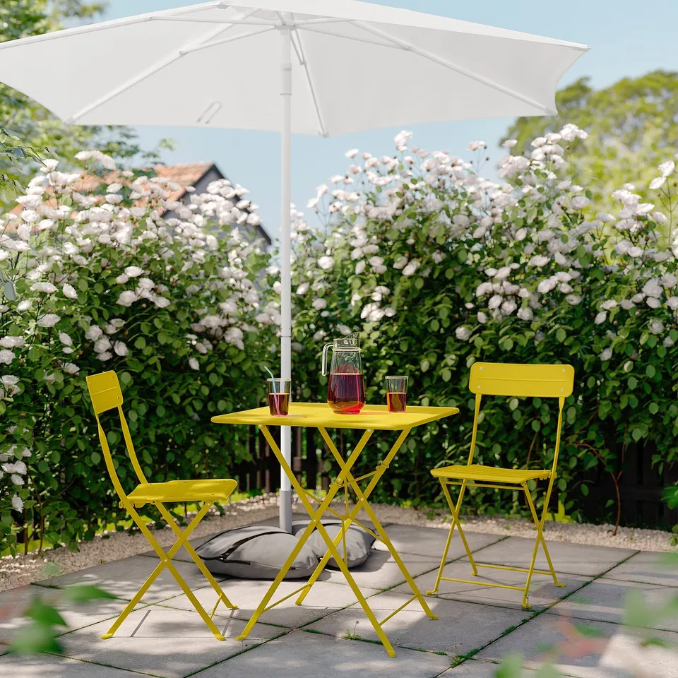Bright yellow metal patio set under white umbrella, surrounded by blooming bushes, includes small table and two folding chairs.