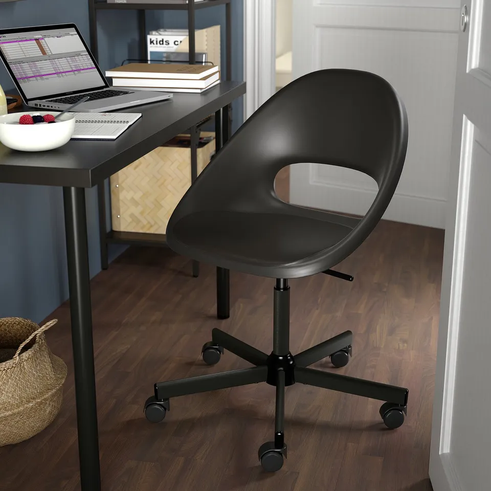 Black ELDBERGET office chair with adjustable height, round shape, and castor wheels in front of a dark desk with a laptop.