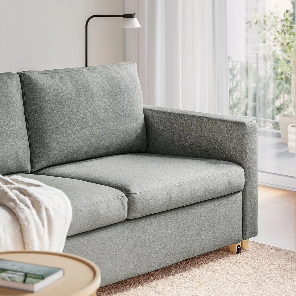 A modern grey sofa-bed with a clean design and comfortable pocket springs, shown with a side table and window in the background.