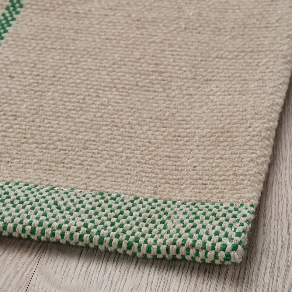 A plush, beige cotton rug with a green chequered border, lying flat on a wooden floor.