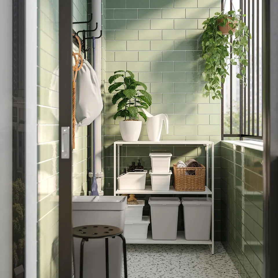 Modern laundry room with green tiles, white fixtures, and a JOSTEIN shelf with plants. Natural light from large window.