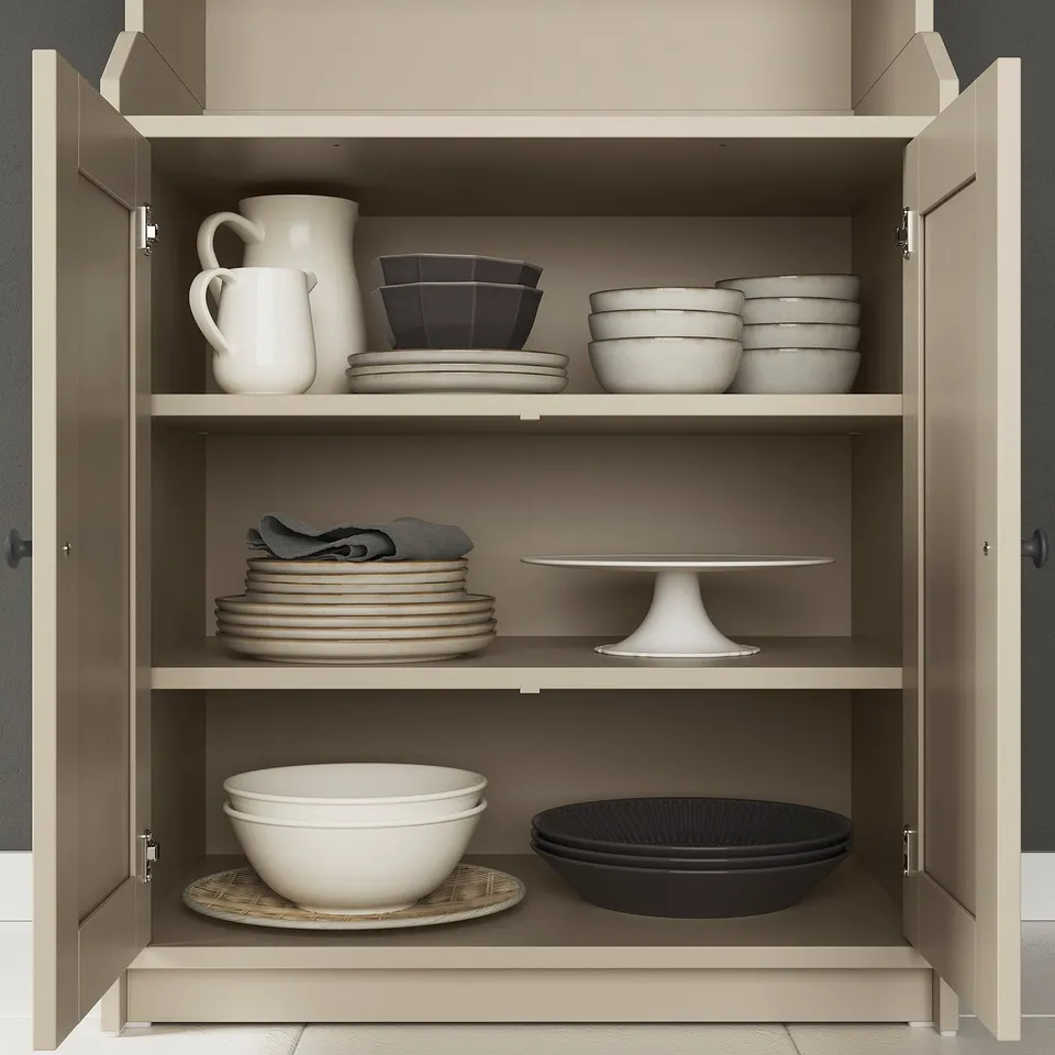 HAUGA beige cabinet with closed doors, storing white and black dishes, bowls, jug, and cake stand.