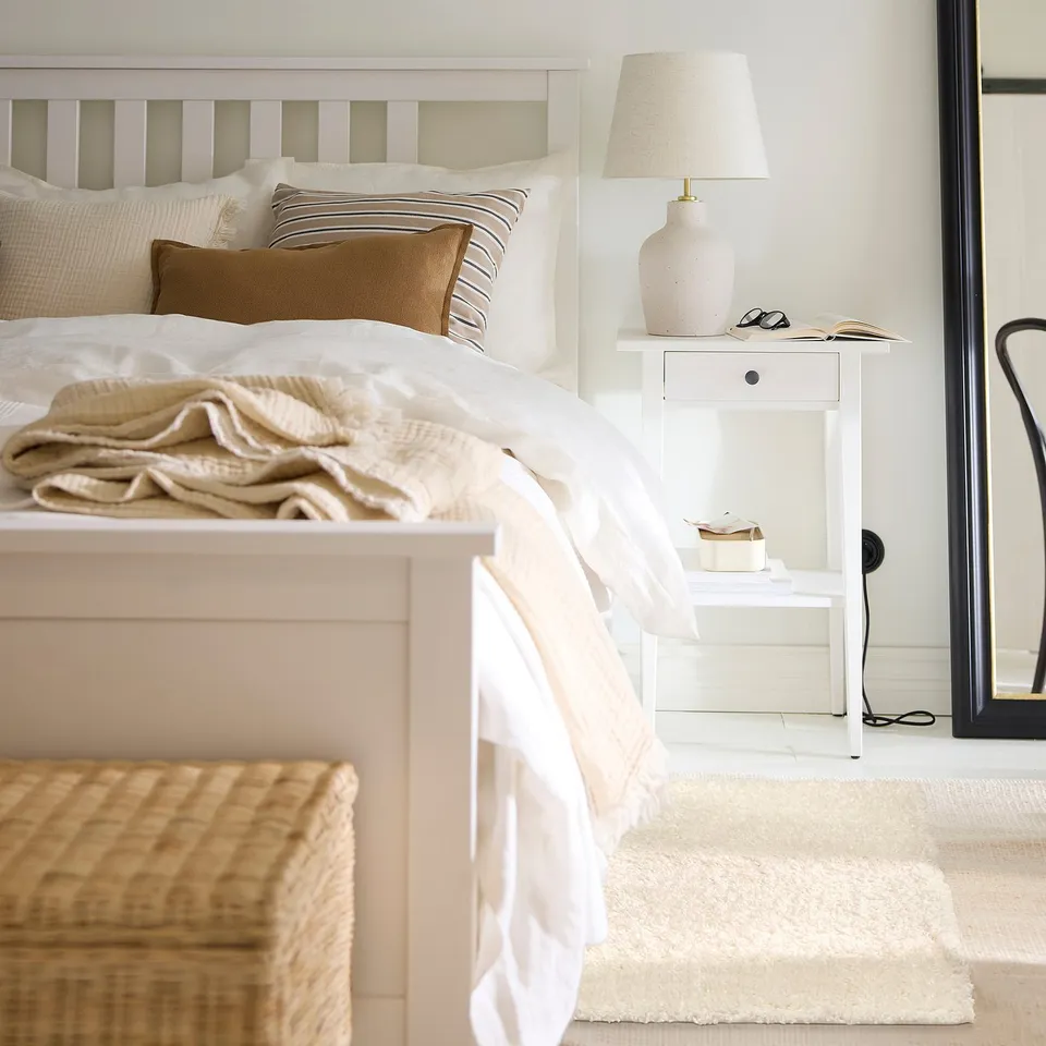 Cosy bedroom with white bedding, sizeable cream-colored ÄRENDE rug on floor, small bedside table with lamp, books, and sunglasses.
