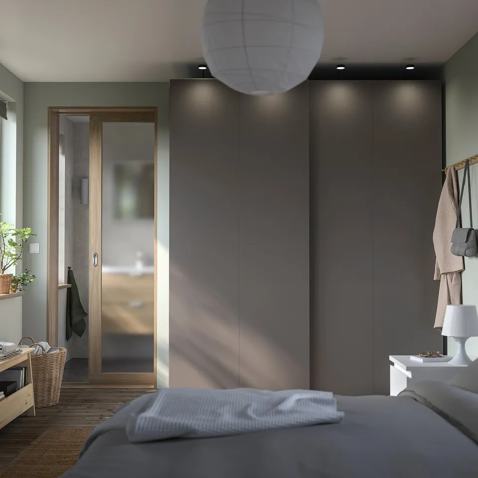 Modern bedroom with grey PAX wardrobe, sliding doors, and white bed linens. Natural light fills the room, complemented by a hanging white lamp.