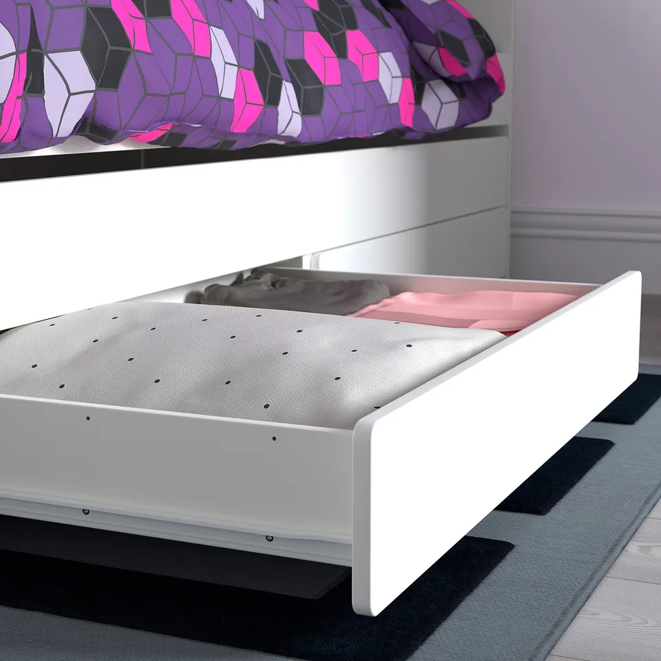 A white bed frame with a lower drawer, part of the SLÄKT series. A cat sleeps on a pink mattress inside the drawer.
