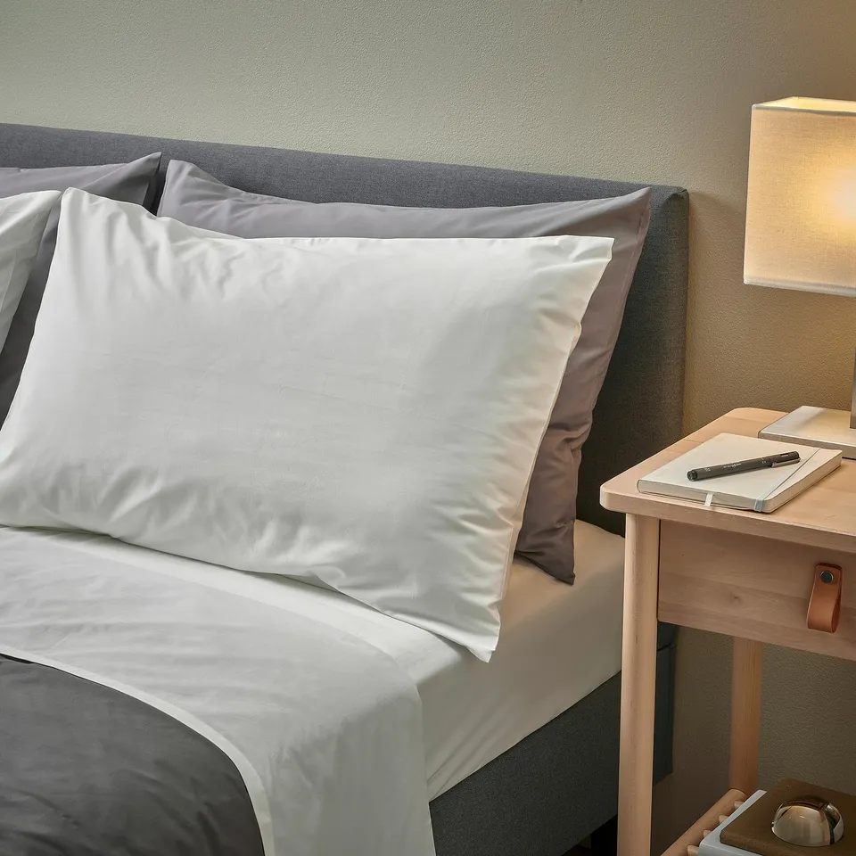 Bed with white RÖNNVECKMAL sheets & pillows, grey headboard, wooden bedside table, lamp, notebook, pen, phone.