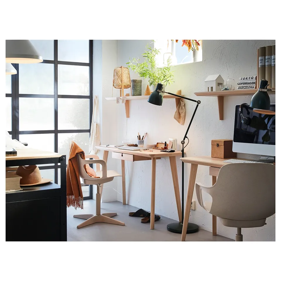 Well-lit home office with natural decor, featuring a light wood LISABO desk and chair, a black table lamp, and shelves with personal items.