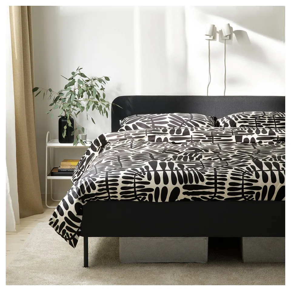 Modern black upholstered bed with padded headboard, abstract print duvet cover, stylish bedside table with plant, beige curtains.