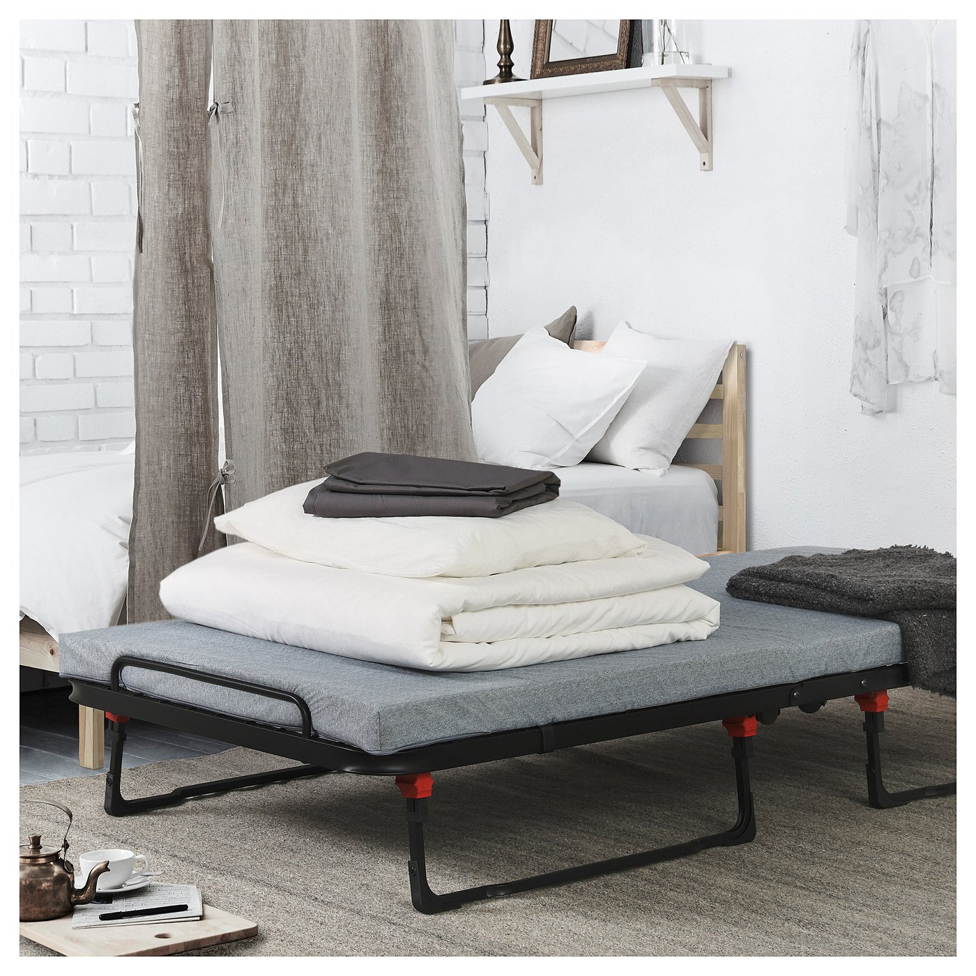 Modern bedroom with white bed, grey fabric guest bed, red feet, folded blankets on top. White curtains, wooden shelf with mirror.
