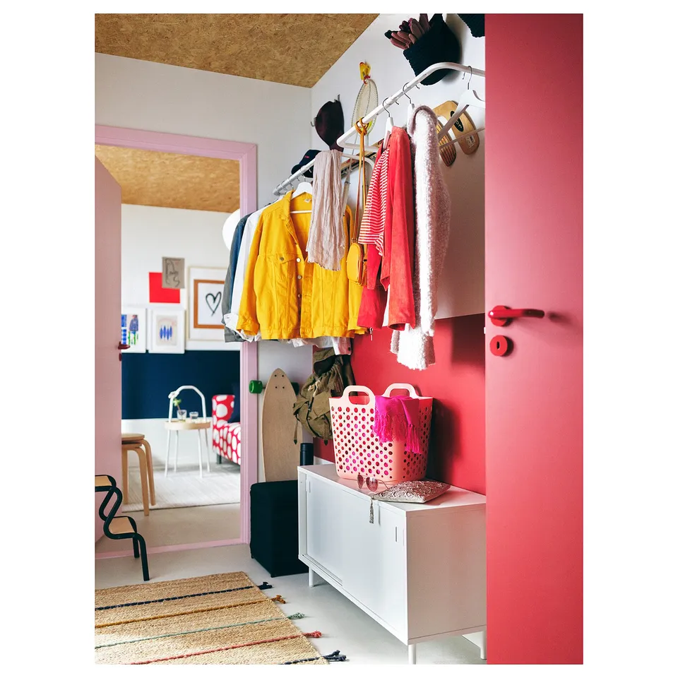 Bright pink room with yellow jackets, pink laundry baskets, white cabinet, colourful rug, and abstract artwork.