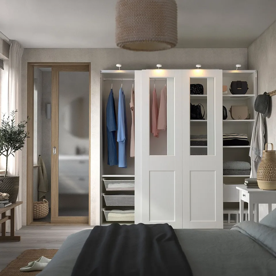 Modern bedroom with large PAX wardrobe, colourful clothes, and warm lamp.