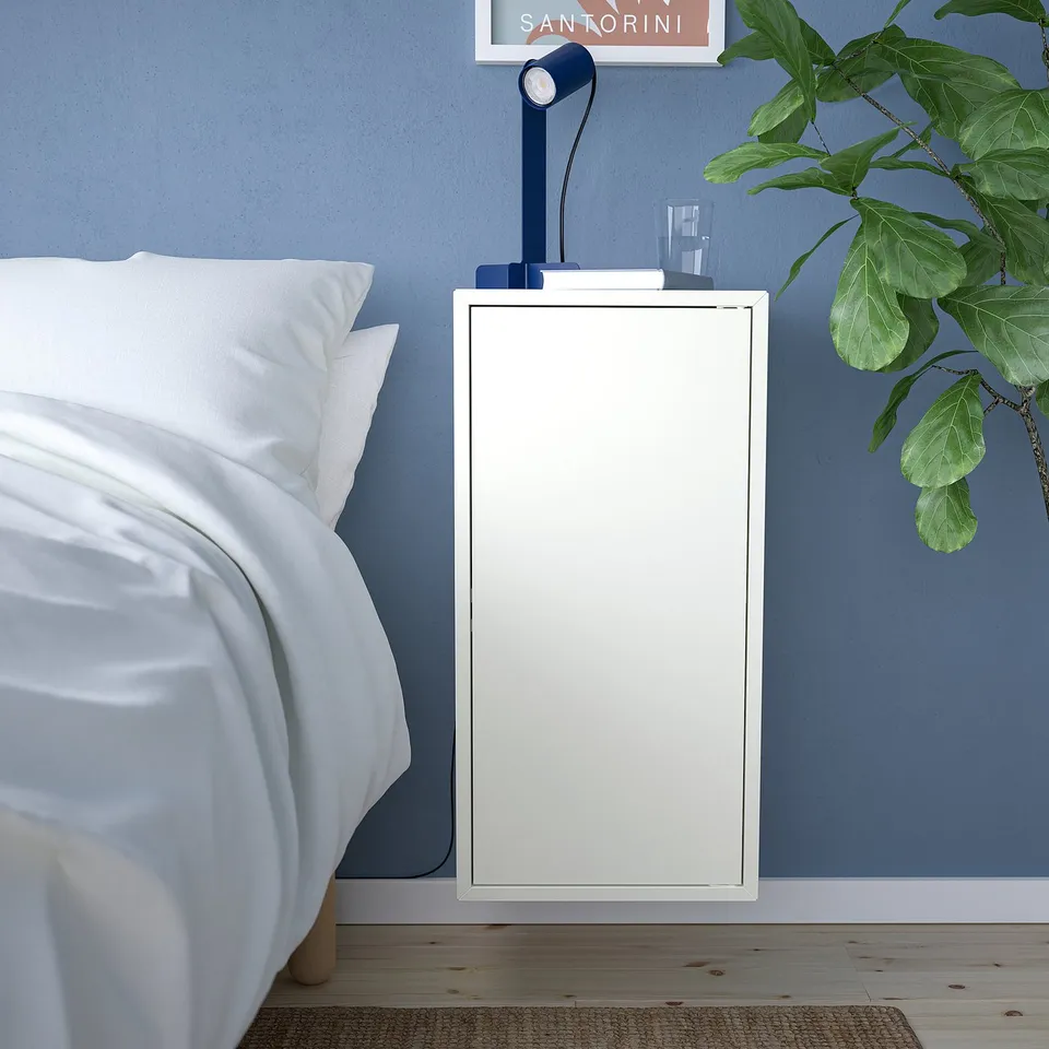 Small white EKET bedside cabinet, slim & tall, next to bed. Holds lamp & book, blue wall behind.
