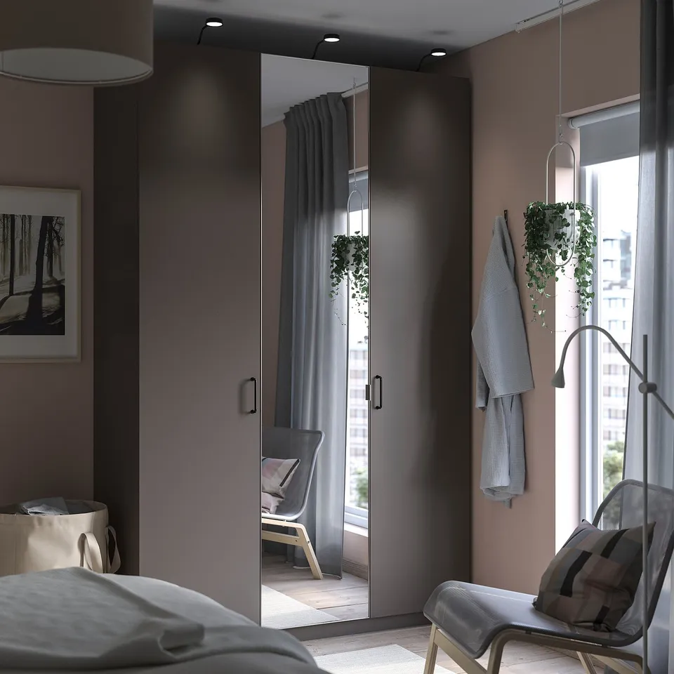 Modern bedroom with grey PAX wardrobe featuring mirror doors, displaying sleek design with hanging plants, and a cosy white chair by the window.