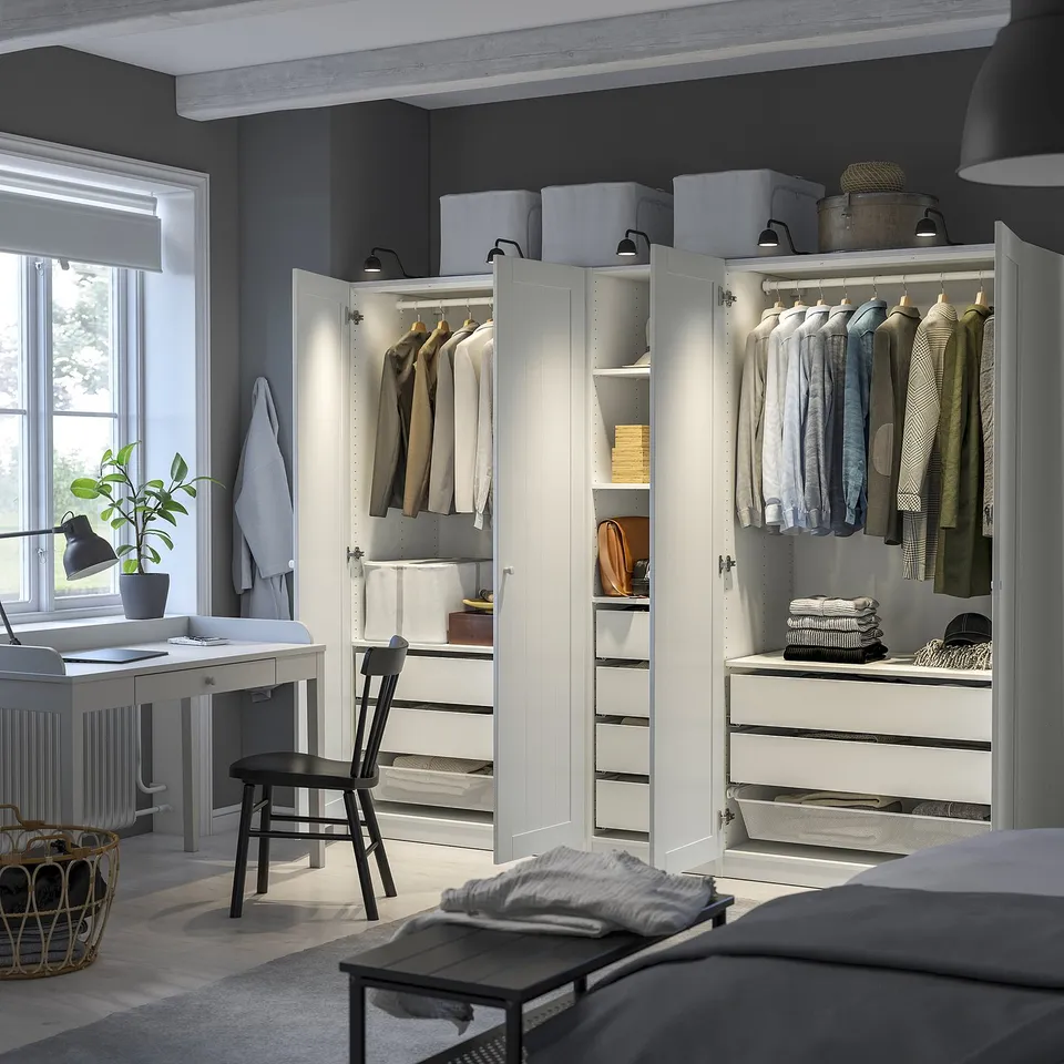 Modern grey wardrobes with lighting, clothes, shelves, drawers, & mesh basket. Nearby, a desk with a chair & plant.