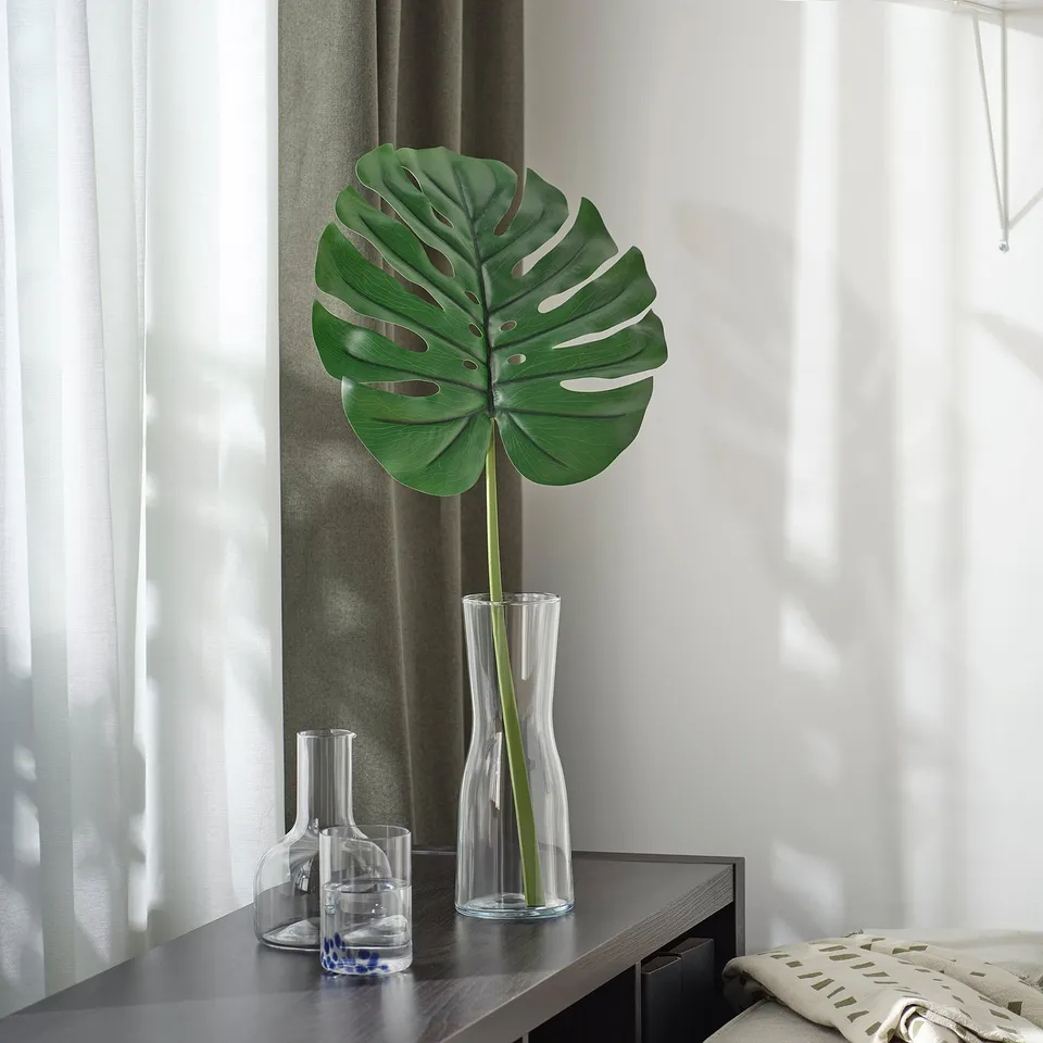 Green artificial leaf in vase, larger than nearby carafes, minimalist setting with light curtains.