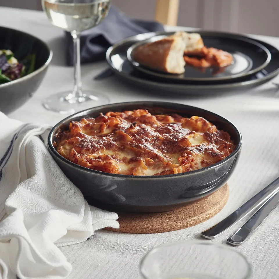Dark oven dish filled with lasagna on table, surrounded by plates, cutlery, and wine glass.
