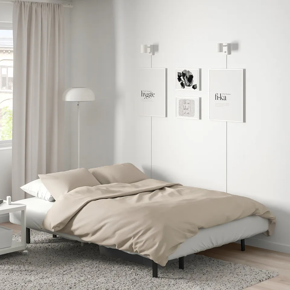A modern bedroom with a NYHAMN sofa-bed in beige, set against a white wall with wall art. Its neutral design complements the minimalistic decor.