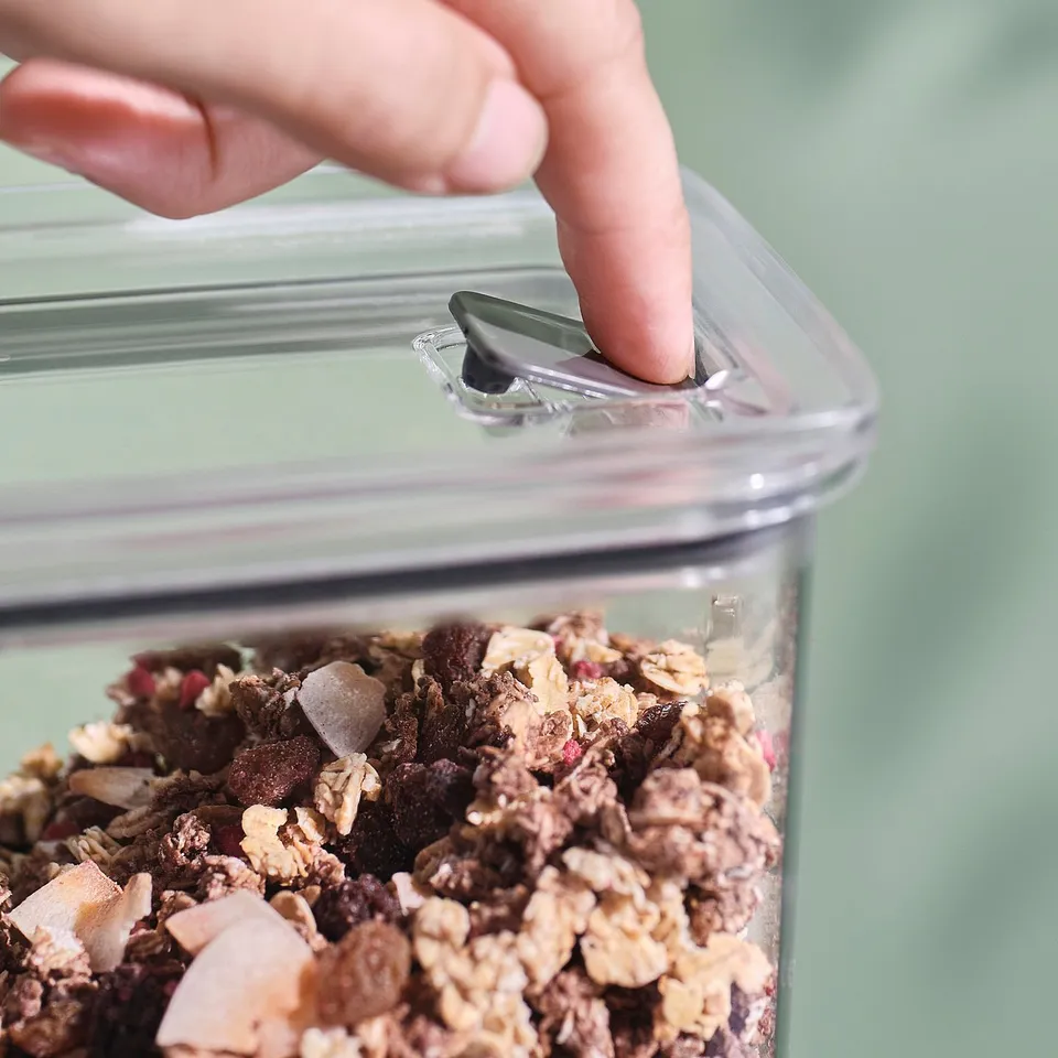 A hand presses a button on a clear, airtight jar lid, releasing air and sealing contents, likely granola, inside.