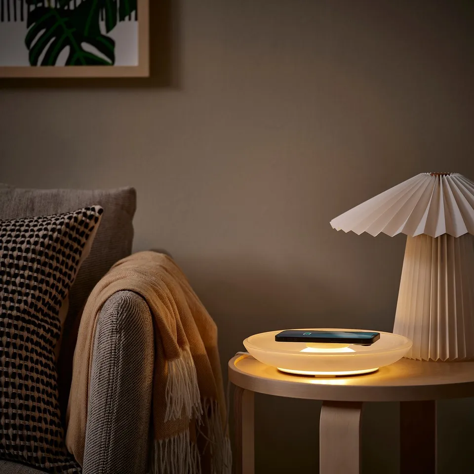 Cosy bedroom scene: side table with VÄSTMÄRKE white wireless charger, soft lamp, and phone. Beside grey pillow and blanket on chair.
