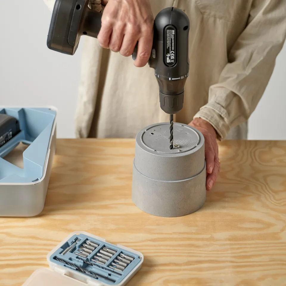 A person drills a concrete-looking planter on a wooden table, demonstrating the products functionality.