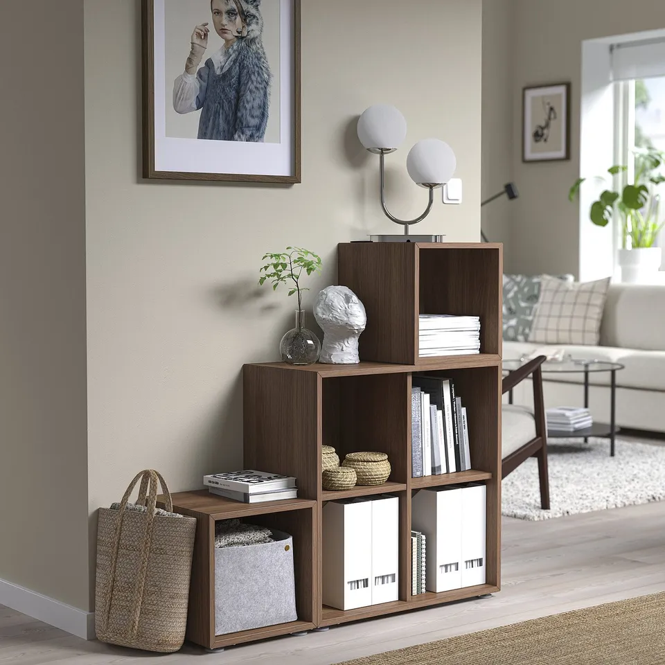 Wooden EKET storage unit with shelves, books, plants, and a chair beside a window. A picture hangs above.